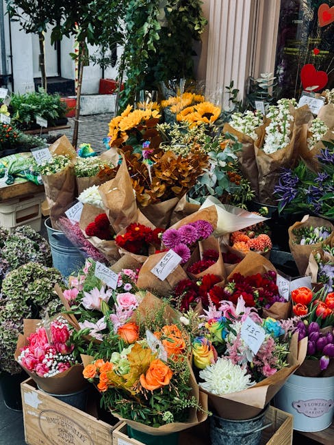 A vibrant display of fresh flower bouquets arranged outdoors on wooden tables and metal buckets, featuring a variety of flowers including bright yellow sunflowers, pink and purple tulips, red and oran