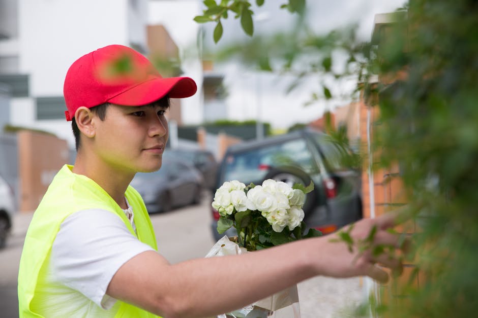 A young male florist from Stamford Hill Florist, wearing a red cap and a high-visibility yellow vest, is arranging a bouquet of fresh white hydrangeas wrapped in brown paper. He stands outdoors on a r