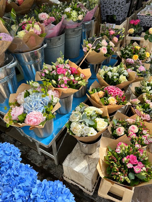 A display of freshly cut flowers arranged in various bouquets and wrapped in brown craft paper or placed in metal buckets. The flowers include pink roses, white and green hydrangeas, light blue hydran