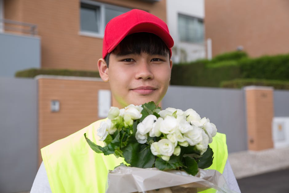 A young male delivery person wearing a red cap and neon yellow vest is holding a bouquet of fresh white flowers, including roses and possibly lisianthus, wrapped in white paper. He stands outdoors in