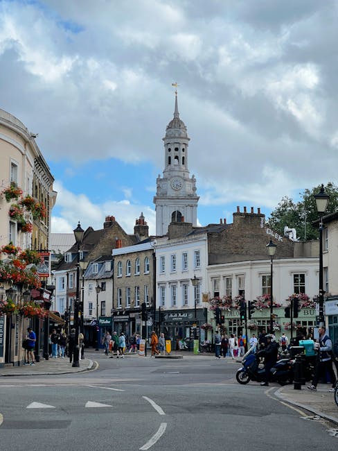The image depicts a busy street scene in a historic town center, with a prominent white clock tower topped by a golden weather vane against a partly cloudy sky. Surrounding the tower are classic Victo