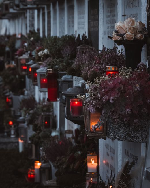 A memorial wall adorned with dozens of illuminated candles and floral arrangements, including roses and chrysanthemums in shades of pink, cream, and white, placed in vases and decorative containers. T