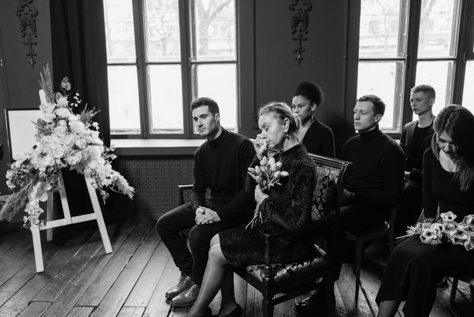 A black-and-white photograph of a funeral scene showing six mourners seated indoors, with a focused view of an older woman holding a bouquet of fresh tulips and roses, dressed in a dark textured coat.