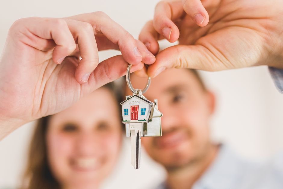 A close-up of a person's hand holding a silver key with a house-shaped keychain featuring a red door, blue windows, and the word 'HOME' inscribed at the top. In the blurred background, a smiling coupl